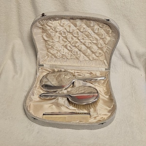 May include: A vanity set in a cream-colored case with a tufted satin interior. The set includes a silver-toned hand mirror, a silver-toned hairbrush with light-colored bristles, and a rectangular cream-colored comb, all nestled in the fabric lining.