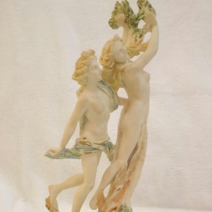 Könnte beinhalten: Eine cremefarbene Skulptur, die zwei Figuren darstellt. Eine Figur hält einen belaubten Zweig über dem Kopf, während die andere zu tanzen scheint. Die Skulptur hat eine klassische Ästhetik mit fließenden Gewändern und detaillierten Gesichtszügen.