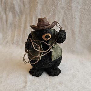 May include: A black bear figurine wearing a brown cowboy hat and a green vest. The bear is holding a coiled tan rope in its paws. The figure has a textured black fur finish, a tan muzzle, and a black nose, standing in an upright position.