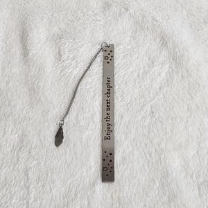 Pode incluir: Um marcador de página de metal prateado com uma barra retangular gravada com o texto "Enjoy the next chapter" rodeado por pequenas estrelas. Uma fina corrente de metal está presa ao topo, terminando num pequeno pingente de pena.