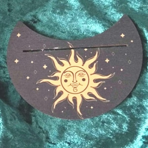 May include: A crescent-shaped black and gold sun and stars design on a wooden coaster. The sun has a smiling face with two eyes and a nose.