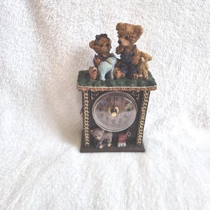 May include: A decorative clock featuring three teddy bear figurines, a lamb, and a small red wagon. The clock has a brown wooden frame with a clear clock face and gold-colored hands. The clock is set on a white surface.