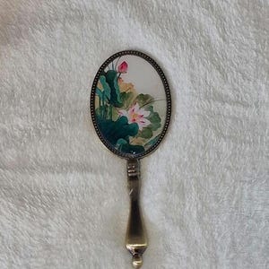 Könnte beinhalten: Ein Vintage-Handspiegel mit einem ovalen Rahmen und einem Messinggriff. Der Spiegel zeigt ein gemaltes Design aus rosa und weißen Lotusblumen mit grünen Blättern auf cremefarbenem Hintergrund.