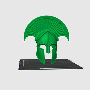 Puede incluir: Un casco espartano verde brillante, con una cresta y protectores de mejillas prominentes. El casco est&aacute; impreso en 3D y descansa sobre una superficie gris oscuro. El dise&ntilde;o es una representaci&oacute;n cl&aacute;sica de los cascos militares griegos antiguos.