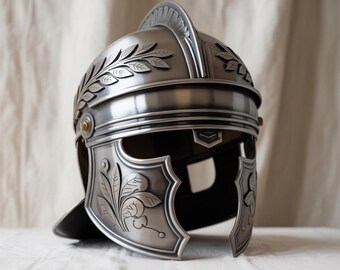 May include: A silver-colored Roman helmet with intricate floral and leaf designs. The helmet features a prominent crest and cheek guards, with a polished finish. The design includes a circular rivet on the side. The helmet is a historical replica.