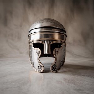May include: A silver-colored Roman helmet with gold-colored trim. The helmet features a detailed design on the top and cheek guards. The helmet has a face guard and is displayed on a marble surface.