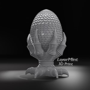May include: A grey 3D-printed dragon egg, held by a claw-like base. The egg has a scale-like pattern. The base has a detailed claw design. The text "LayerMint 3D Print" is visible on the base.