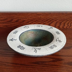 May include: A round, off-white ceramic bowl with a crackled finish. The rim features black symbols and the interior has a metallic green and blue glaze. The bowl sits on a dark wooden surface.