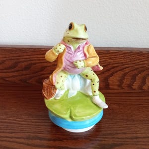 May include: A ceramic figurine of a frog dressed in a yellow jacket and pink vest, sitting on a green lily pad base. The frog has brown spots on its limbs and is holding a small brown woven basket at its side. The base is blue and white.
