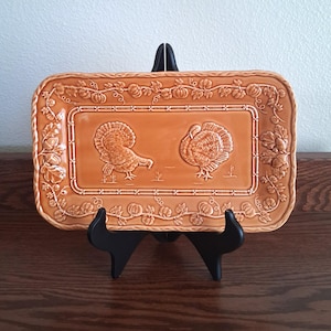 May include: A rectangular orange ceramic serving platter featuring two embossed turkeys in the center. The border is decorated with a raised relief pattern of pumpkins, leaves, and acorns. The platter is displayed on a black plastic stand.