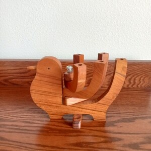 May include: A wooden bird-shaped ring holder with a natural wood grain finish. The holder features a bird body, a beak, and curved supports to hold rings. The ring holder is displayed on a wooden surface.