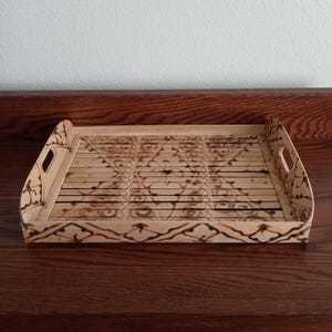 May include: A rectangular wooden serving tray with a decorative pattern. The tray has curved handles and a light brown color with darker brown accents. The design features a series of lines and geometric shapes.