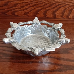 May include: A small, silver-colored decorative dish with a detailed design. The dish features a textured interior and is adorned with sculpted animal figures around the rim. The dish is made of metal and sits on a wooden surface.