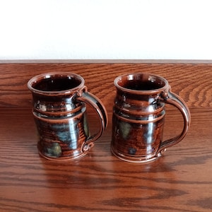 May include: Two handmade ceramic mugs with a dark brown glaze and a textured surface. The mugs have a cylindrical shape with a handle and a slightly flared rim. The glaze has hints of blue and green. The mugs are displayed on a wooden surface.