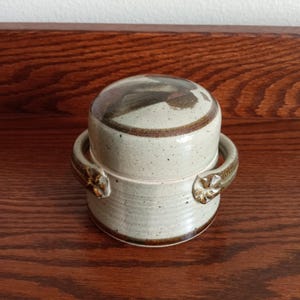 May include: A speckled beige ceramic lidded jar with two side handles featuring small flower-shaped accents. The jar has a cylindrical body with horizontal ridges and a smooth, rounded lid with dark brown and grey abstract glaze patterns.