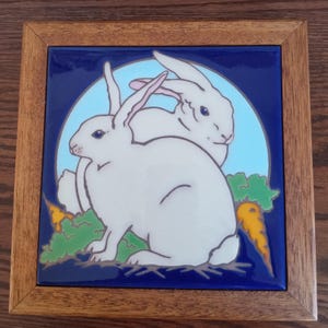 May include: Square ceramic tile with a blue background and a light blue circle featuring two white rabbits. The rabbits are sitting near green leaves and orange carrots. The tile is set in a brown wooden frame.