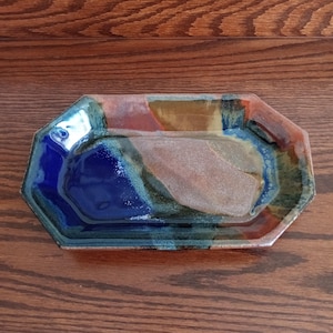 May include: An octagonal ceramic tray featuring a multi-colored glaze with deep blue, earthy brown, and muted green tones. The surface displays a glossy finish with a textured, speckled central area that transitions into smooth, vibrant color gradients.