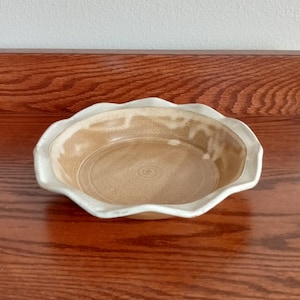 May include: A handmade ceramic pie dish with a scalloped edge. The dish has a light brown interior and a creamy white rim. The pie dish is designed for baking and serving pies.