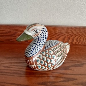May include: A ceramic duck figurine with a detailed, hand-painted design. The duck features a white body with brown, blue, and green accents. The duck's head has a speckled pattern, and the beak is green.