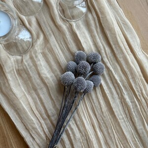 May include: A cluster of gray, spherical decorative flowers with dark stems tied with twine. The flowers rest on a beige, textured fabric, with three clear glass candle holders with lit white candles in the background.