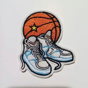 Può includere: Toppa ricamata raffigurante un pallone da basket e scarpe da ginnastica. Il pallone è arancione con una stella gialla. Le scarpe da ginnastica sono bianche con dettagli azzurri e lacci neri. La toppa ha un bordo nero.