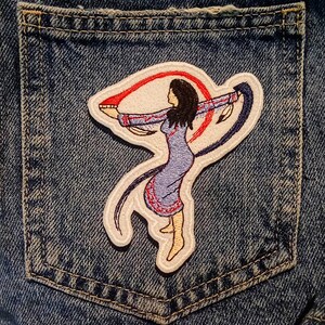 May include: Embroidered patch depicting a Native American woman in a blue dress, arms outstretched, holding red and blue ribbons. The patch is on a denim pocket, with detailed stitching.
