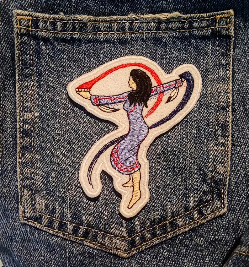 May include: Embroidered patch depicting a Native American woman in a blue dress, arms outstretched, holding red and blue ribbons. The patch is on a denim pocket, with detailed stitching.