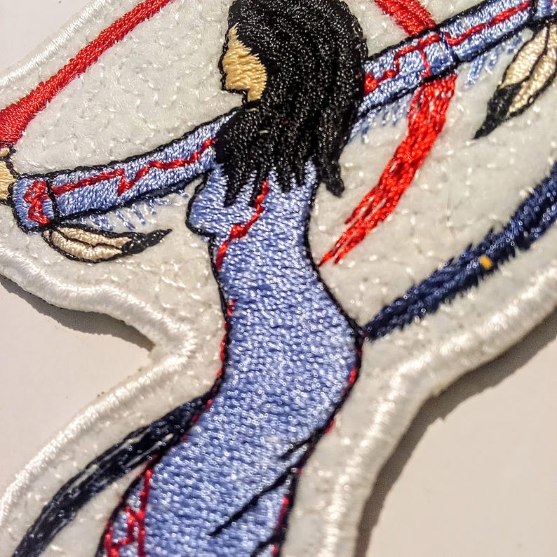 May include: Embroidered patch depicting a Native American woman in a blue dress with red and blue detailing. The patch has a white background, showcasing the woman's silhouette, long black hair, and outstretched arms. Feathers and decorative elements are also included in the design.