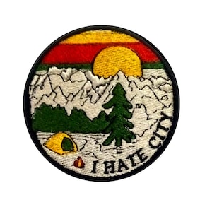 Può includere: Una toppa rotonda ricamata con una scena di montagna, un sole giallo, un albero verde e una tenda gialla. Il testo "I HATE CITY" è scritto in un cerchio nero attorno alla scena.