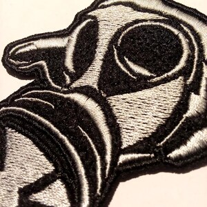 May include: Close-up of a black and white embroidered patch. The patch features a stylized design with rounded shapes and curved lines. The design is primarily black with white outlining, creating a high-contrast effect. The patch has a textured appearance.