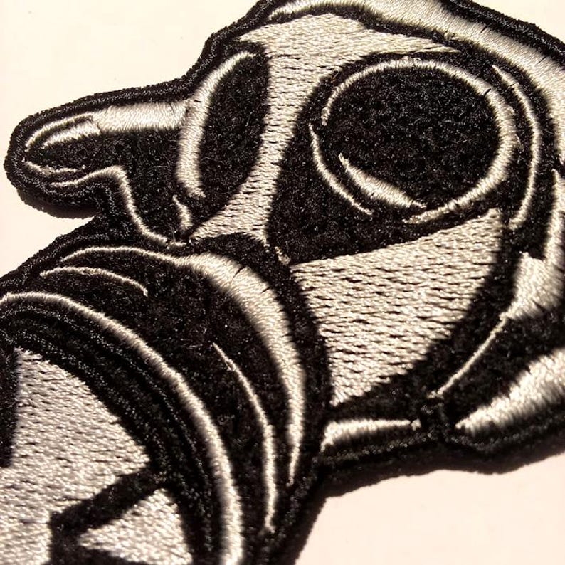 May include: Close-up of a black and white embroidered patch. The patch features a stylized design with rounded shapes and curved lines. The design is primarily black with white outlining, creating a high-contrast effect. The patch has a textured appearance.