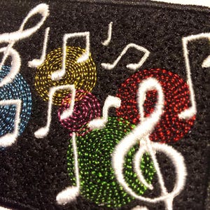 May include: Close-up of a black fabric embroidered with white musical notes and colorful, textured circles. The circles are in shades of blue, yellow, red, green, and purple. The musical notes are in white.