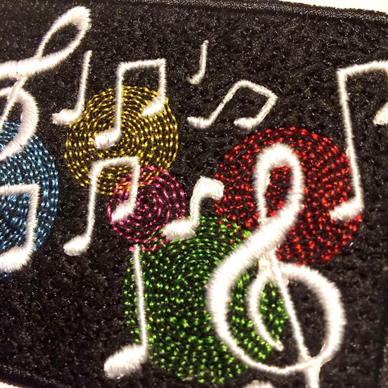 May include: Close-up of a black fabric embroidered with white musical notes and colorful, textured circles. The circles are in shades of blue, yellow, red, green, and purple. The musical notes are in white.