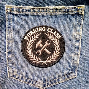 May include: A denim pocket in blue, with a circular patch stitched on. The patch reads "WORKING CLASS" in white, framing a crossed hammer and spanner design. A white laurel wreath surrounds the tools on a black backdrop.