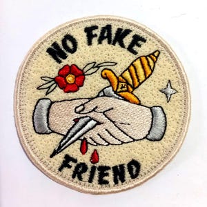 Può includere: Una toppa ovale beige ricamata con il testo "NO FAKE FRIEND" in nero. Un'immagine stilizzata di una stretta di mano con un pugnale che trafigge le mani e una goccia di sangue. Un fiore rosso con foglie verdi si trova nell'angolo in alto a sinistra.