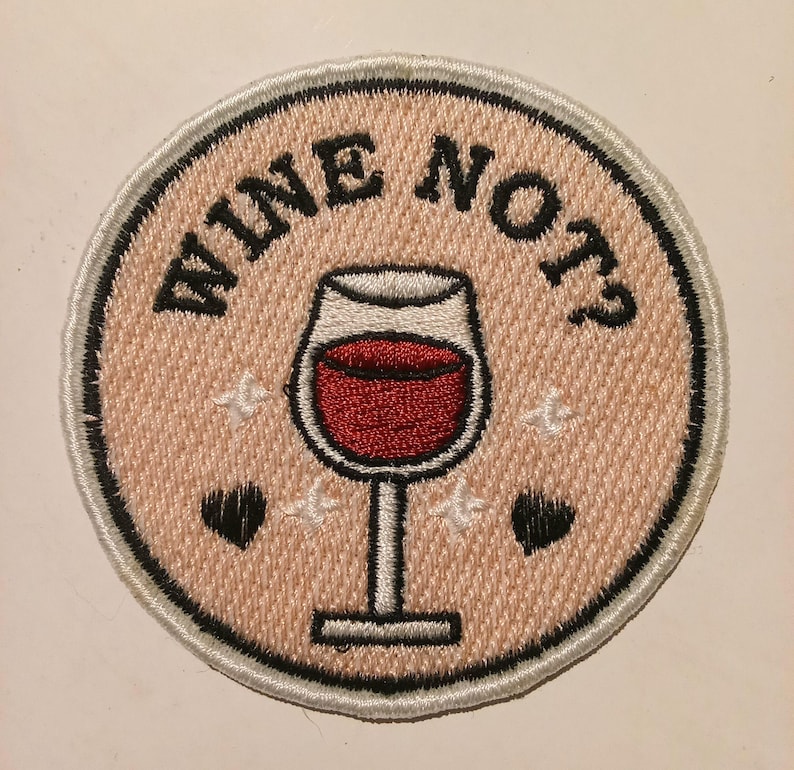 May include: Embroidered patch featuring the text "WINE NOT?" in black, encircling a wine glass filled with red liquid. The patch has a pale pink backdrop with white stars and black hearts.