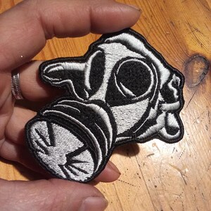 May include: Embroidered patch featuring a stylized gas mask design. The patch is primarily black and white, with detailed stitching outlining the mask's features. The patch is held in a hand, showcasing its size and shape.