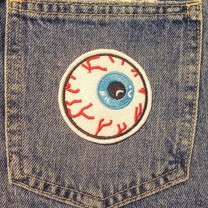 May include: A circular embroidered patch with an eyeball design. The eye has a blue iris, a black pupil, and red veins on a white background. The patch is sewn onto the pocket of a blue denim garment.