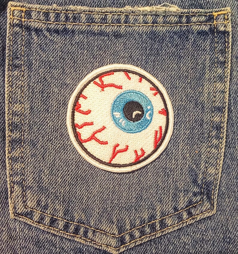 May include: A circular embroidered patch with an eyeball design. The eye has a blue iris, a black pupil, and red veins on a white background. The patch is sewn onto the pocket of a blue denim garment.