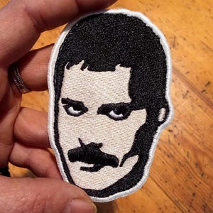 Op de afbeelding: Geborduurde patch met een zwart-wit portret van een man met een opvallende snor en kapsel. De patch heeft een witte rand en is ontworpen om op stof te strijken of te naaien. Het ontwerp is een gestileerde weergave.