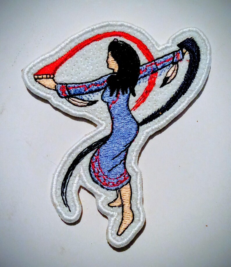 May include: Embroidered patch showing a woman in a blue dress with red and black details, arms extended. The patch has a white border and depicts a stylized figure with long black hair. The design includes a red arc and a black arc.