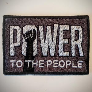 Può includere: Toppa ricamata con le parole "POWER TO THE PEOPLE" in filo bianco su sfondo rettangolare grigio scuro. Un pugno nero è raffigurato al centro. La toppa ha un bordo nero.