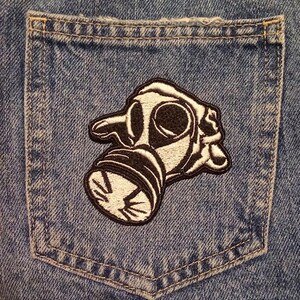 May include: A denim pocket with a black and white embroidered patch of a gas mask. The patch is detailed with black outlines and white fill, and is sewn onto the blue denim fabric.