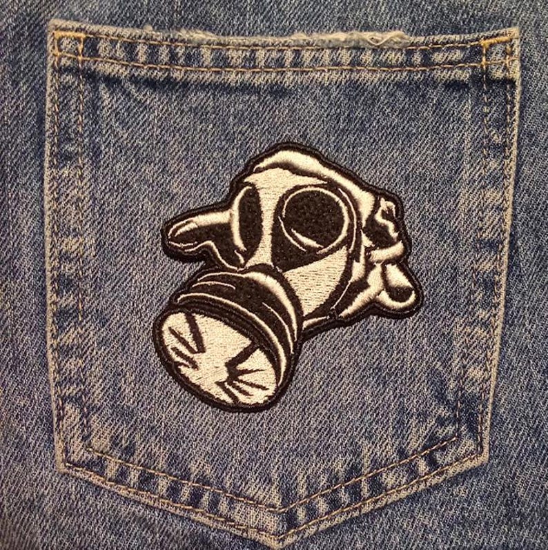 May include: A denim pocket with a black and white embroidered patch of a gas mask. The patch is detailed with black outlines and white fill, and is sewn onto the blue denim fabric.