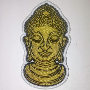 Può includere: Toppa ricamata raffigurante una testa di Buddha dorata con occhi chiusi e un'espressione serena. La toppa ha un bordo bianco con un contorno blu navy. Adatta per abbigliamento o accessori.