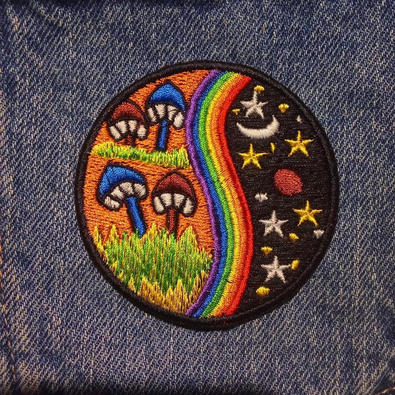 Peut inclure: &Eacute;cusson brod&eacute; circulaire avec une bordure noire. Le motif comprend des champignons, un arc-en-ciel, des &eacute;toiles et un croissant de lune. L'&eacute;cusson est cousu sur un tissu en jean bleu. Le motif de l'&eacute;cusson est divis&eacute; en deux.