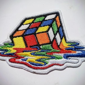 Peut inclure: Écusson brodé représentant un Rubik's Cube en train de fondre. Les carrés du cube sont de couleurs vives : rouge, orange, jaune, vert, bleu et blanc. Le patch a une bordure blanche et un effet dégoulinant coloré.