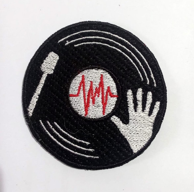 Może przedstawiać: Czarno-biała haftowana łatka przedstawiająca płytę winylową z czerwonym wzorem bicia serca i białą ręką sięgającą do płyty.