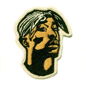 May include: Embroidered patch depicting a portrait of a man wearing a bandana. The patch is in black and gold, with a white border. The design is a stylized representation of the subject's face.