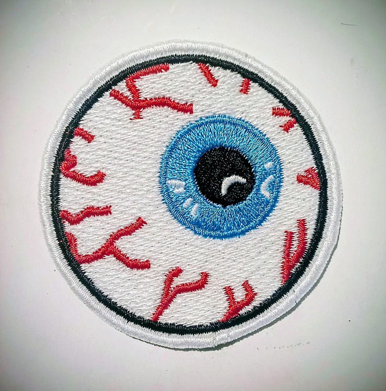 May include: Embroidered patch with an eyeball design. The eye has a bright blue iris, a black pupil, and red veins on a white background. The patch is round with a black border, suitable for customising clothing or accessories.